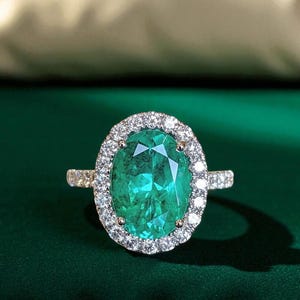 Puede incluir: Anillo con esmeralda de talla ovalada rodeada de pequeños diamantes. La banda del anillo también está adornada con pequeños diamantes. La esmeralda es de color verde vibrante y los diamantes brillan sobre un fondo verde oscuro.
