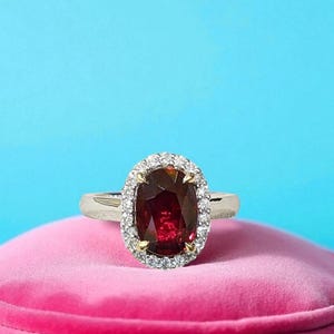 Puede incluir: Un anillo con una gran piedra preciosa de color rojo intenso y corte ovalado, rodeada por un halo de pequeñas piedras blancas transparentes. La piedra central está sujeta por cuatro garras de oro amarillo sobre una banda de metal plateado pulido.