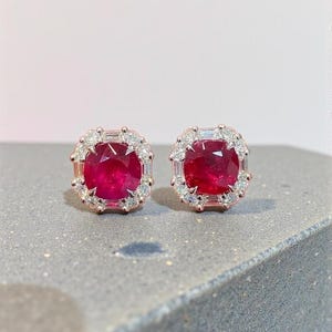May include: A pair of stud earrings featuring a central cushion-cut deep red ruby surrounded by a halo of small, clear, baguette and round-cut diamonds set in a rose gold metal frame. The earrings have a square shape with rounded corners.