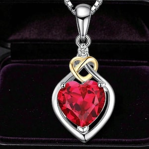 Può includere: Una collana con pendente color argento caratterizzata da una grande pietra rossa a forma di cuore incastonata in una cornice d'argento. Sopra il cuore rosso c'è un piccolo design a cuore intrecciato dorato con una pietra chiara sulla maglia.