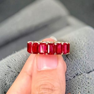 Può includere: Una fede in oro con una fila continua di gemme rosso vivo dal taglio a smeraldo, montate su una struttura a griffe condivise. L'anello presenta una serie di pietre rettangolari che circondano la fascia, mostrando profonde tonalità cremisi.
