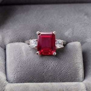 Op de afbeelding: Een ring met een grote, rechthoekige, smaragdgeslepen rode edelsteen in een gepolijste metalen band. De middelste steen wordt geflankeerd door twee kleinere, ronde, heldere gefacetteerde stenen, elk vastgezet met een zetting van vier pootjes.