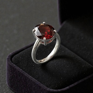 Op de afbeelding: Een zilveren ring met een grote, ovaal geslepen, dieprode granaat edelsteen in een zetting met vier pootjes. De ringband is glad en gepolijst en vertoont een metaalachtige glans die contrasteert met de gefacetteerde steen in het midden.