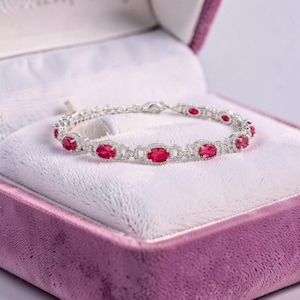 Puede incluir: Una pulsera de eslabones en tono plateado que presenta una serie de piedras preciosas de color rojo vibrante con corte ovalado. Cada piedra está engastada en un marco metálico decorativo, unidas por pequeños eslabones circulares.