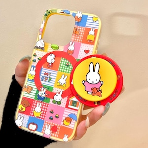 Op de afbeelding: Een gele telefoonhoes met een kleurrijk patchwork-ontwerp met cartoonkonijntjes en andere figuren. Een bijpassende popsocket met een konijn en een hond is aan de hoes bevestigd. De popsocket is rood en geel.