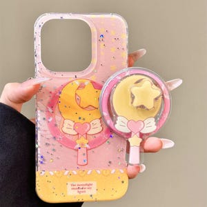 Puede incluir: Funda de teléfono rosa con un diseño brillante, con un motivo de luna y estrella amarilla con un corazón y un lazo rosa. La funda tiene un dije a juego. El texto en la funda dice "The moonlight stands for my heart."