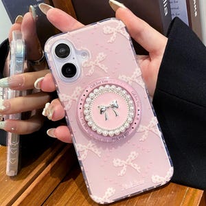 以下が含まれることがあります： ピンク色の携帯電話ケース。リボン模様と円形のグリップが付いています。グリップはパールとシルバーのリボンで飾られています。ケースには「Gift For You」と「Pink Bow」の文字がプリントされています。