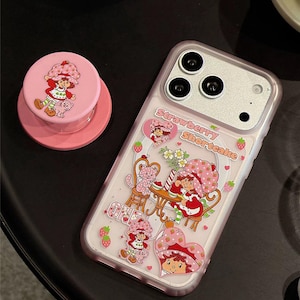 Könnte beinhalten: Rosa Handyhülle und passender Pop-Socket mit Erdbeer-Schortcake-Illustrationen. Die Hülle hat einen transparenten Hintergrund mit Erdbeer-Schortcake-Figuren. Der Pop-Socket hat eine rosa Basis und ein Erdbeer-Schortcake-Design. Der Text "Strawberry Shortcake" ist sichtbar.
