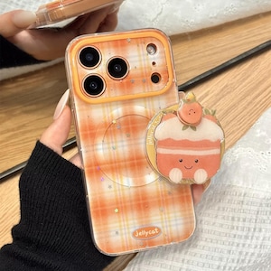 Könnte beinhalten: Transparente Handyhülle mit orangefarbenem Karomuster und einem cupcakeförmigen Handyhalter. Die Hülle hat einen transparenten Rand und ein Jellycat-Logo. Die Handyhülle wird in der Hand einer Person gehalten.