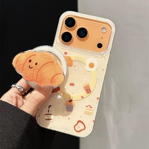 Puede incluir: Funda de teléfono con fondo amarillo claro e ilustraciones de temática alimentaria. La funda tiene un agarre desplegable con un diseño de croissant sonriente. Otras ilustraciones incluyen un huevo frito, una magdalena y una taza de café.
