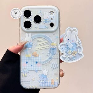 May include: A clear phone case with a blue and white bunny pattern, featuring illustrations of bunnies, stars, and text. A matching bunny-shaped phone grip is attached. The case has a protective bumper around the camera.