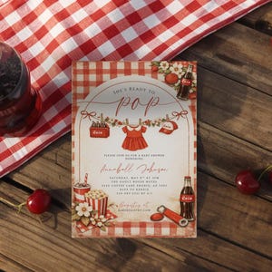 May include: Baby shower invitation with a red and white gingham pattern. The invitation features the text "She's Ready to Pop" and includes a red dress graphic. A bottle of cola, cherries, and popcorn are also visible.