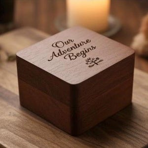 Könnte beinhalten: Eine quadratische Holzbox in Dunkelbraun mit einem Klappdeckel. Auf dem Deckel sind die Worte "Our Adventure Begins" in einer dekorativen Schriftart und ein kleines Baum-Symbol eingraviert. Die Box steht auf einer Holzoberfläche.