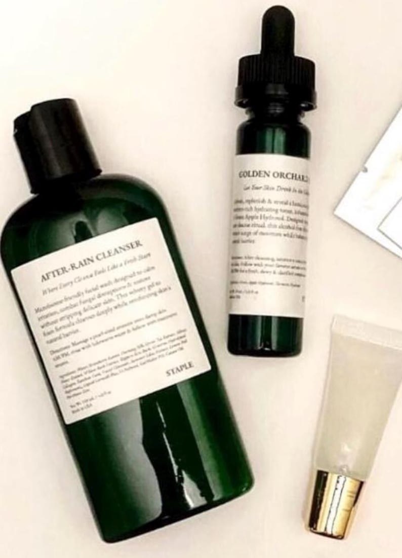 Pode incluir: Uma cole&ccedil;&atilde;o de produtos para a pele, incluindo um frasco verde escuro de "AFTER-RAIN CLEANSER" e um frasco pequeno com o r&oacute;tulo "GOLDEN ORCHARD". Um tubo transparente com tampa dourada tamb&eacute;m est&aacute; presente. Sobre uma superf&iacute;cie branca.