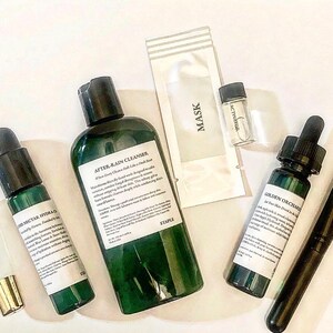 Pode incluir: Uma cole&ccedil;&atilde;o de produtos para a pele, incluindo um frasco verde rotulado "AFTER-RAIN CLEANSER", um pequeno frasco de "ACTIVATOR", uma m&aacute;scara facial e um pincel. Os produtos est&atilde;o dispostos sobre uma superf&iacute;cie branca.