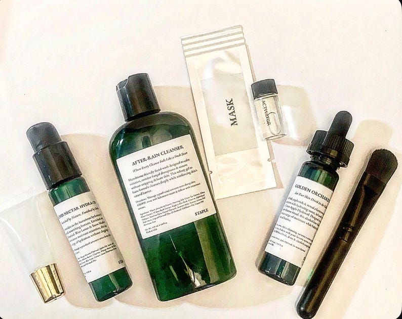 Pode incluir: Uma cole&ccedil;&atilde;o de produtos para a pele, incluindo um frasco verde rotulado "AFTER-RAIN CLEANSER", um pequeno frasco de "ACTIVATOR", uma m&aacute;scara facial e um pincel. Os produtos est&atilde;o dispostos sobre uma superf&iacute;cie branca.