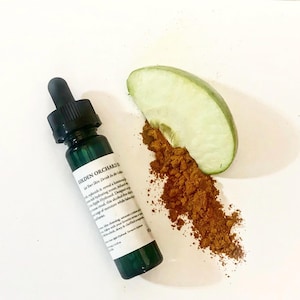 May include: A green glass bottle with a dropper, labelled "Golden Orchard", beside a slice of green apple and a pile of brown powder. The bottle is on a white surface, suggesting a skincare or beauty product.