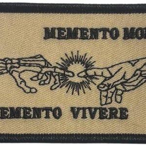 Puede incluir: Un parche rectangular bordado con fondo beige y borde negro. Presenta una ilustración de líneas negras de una mano esquelética alcanzando una mano humana, con un diseño de rayos de sol entre ellas. El texto MEMENTO MORI y MEMENTO VIVERE está bordado en negro.
