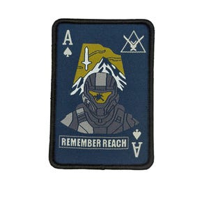 May include: A navy blue patch with a black border, featuring a stylized illustration of a soldier's helmet and the text "REMEMBER REACH." The design includes the Ace of Spades and a mountain scene with a sword.