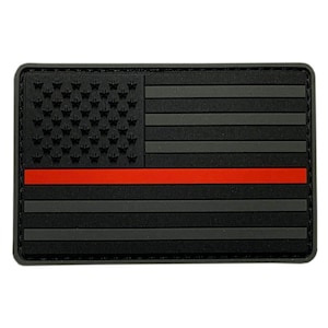 May include: A rectangular morale patch featuring a stylized American flag design in monochromatic black and dark gray tones. The design includes a single horizontal red stripe across the center, with raised stars in the canton area.