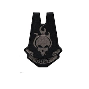 May include: A black embroidered patch featuring a stylized gray skull design with flames above it. Below the skull, a banner contains the text ODST. The patch has a distinct shield-like shape with a notched top edge and dark stitching.