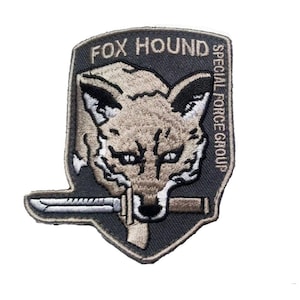May include: A shield-shaped embroidered patch with a dark grey background featuring a tan fox head with black details. A silver knife is held in the fox's mouth. The text FOX HOUND is at the top and SPECIAL FORCE GROUP is on the right side.