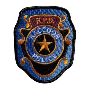 May include: A shield-shaped embroidered patch featuring a blue background with a central gold star. The text R.P.D. is at the top, and the words RACCOON POLICE are arched around the star. The design includes gold scrollwork and a black border.