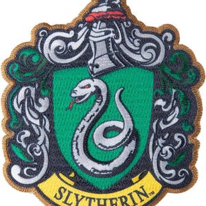 Puede incluir: Un parche bordado que presenta un escudo verde con una serpiente plateada, rematado con un casco plateado y un detalle rojo. El diseño incluye volutas plateadas, un borde dorado y una cinta amarilla en la parte inferior con el texto SLYTHERIN.