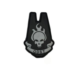 Könnte beinhalten: Schwarzer und grauer ODST-Aufnäher mit einem Totenkopf und Flammen sowie dem Text "ODST". Der Aufnäher hat eine einzigartige Form mit einem Ausschnitt oben. Das Design ist ein stilisiertes Emblem.