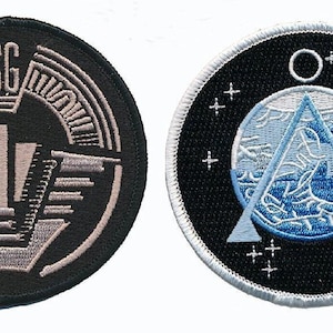 May include: Two circular embroidered patches. The left patch is black with silver-toned thread forming the letters SG, the number 1, and geometric lines. The right patch is black with a white border, featuring a blue triangle and star symbols.