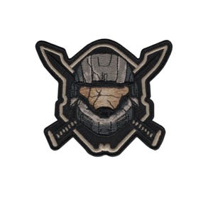 May include: Embroidered patch featuring a stylized helmet design with crossed swords. The patch has a black border with tan and gray details, and a textured appearance. The design is centered on a white background.