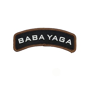 Puede incluir: Un parche negro arqueado con el texto blanco "BABA YAGA". El parche tiene un borde marrón. Es probable que sea un parche bordado o tejido, diseñado para coser o planchar en prendas o accesorios.