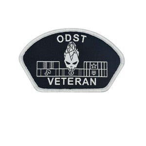 May include: Black and white embroidered patch with the text "ODST VETERAN". The patch features a stylized skull design and various symbols. The patch has a white border and is in a shield shape.