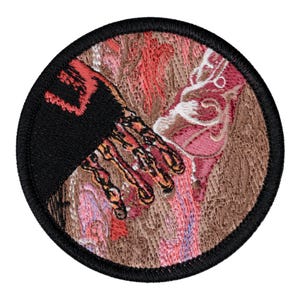 May include: A circular embroidered patch featuring a black sleeve and a hand with orange and yellow skeletal details reaching toward a textured, swirling background of pink, brown, and white thread. The patch has a thick black stitched border.