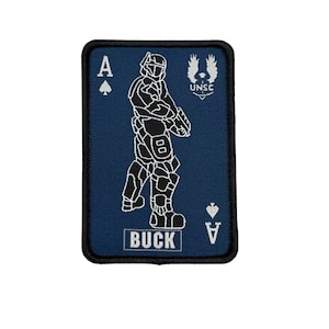 May include: A navy blue patch with a black border featuring a white outline of a soldier holding a weapon. The patch has the text "BUCK" and the Ace of Spades and Ace of Diamonds symbols. The text "UNSC" is also present.