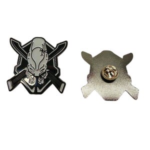 May include: A metal enamel pin featuring a white, stylised helmet design centred over two crossed black swords against a black and white shield background. The reverse side shows a silver-toned metal finish with a circular pin backing mechanism.
