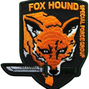 May include: A shield-shaped embroidered patch with a black background featuring an orange fox head with white accents. The text FOX HOUND is stitched in orange at the top, and SPECIAL FORCE GROUP is stitched vertically in orange on the right side.