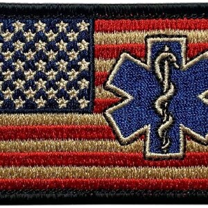 May include: A rectangular embroidered patch featuring the United States flag with red, tan, and dark blue threads. A blue Star of Life symbol with a white embroidered serpent and staff is centered on the right side over the stripes.