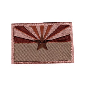 May include: A rectangular embroidered patch featuring a stylized sunburst design in shades of dusty pink and dark brown. A central five-pointed star is positioned over the lower half, which is a solid tan color. The entire patch has a stitched border.