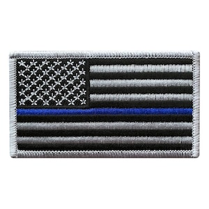May include: A rectangular embroidered patch featuring a stylized American flag design. The flag consists of horizontal black and grey stripes with a single horizontal blue stripe, and a black canton filled with white embroidered stars.