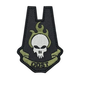 May include: Black embroidered patch with a white skull, olive green flames and accents. The patch has a banner with the text "ODST" in olive green. The patch is shaped like a stylized emblem.