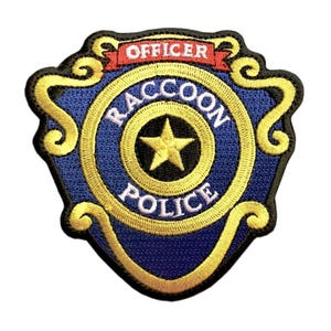 May include: A shield-shaped embroidered patch featuring a blue centre with a yellow star, surrounded by a yellow border. The text reads OFFICER in red on a banner, and RACCOON POLICE in white lettering encircles the central yellow star.