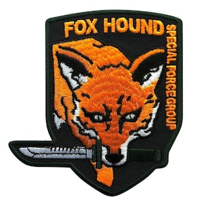 Op de afbeelding: Een schildvormige geborduurde patch met een zwarte achtergrond met een oranje vossenkop en een zilverkleurig mes in de bek. De tekst FOX HOUND is in oranje bovenaan geborduurd, met SPECIAL FORCE GROUP verticaal geschreven aan de rechterkant.