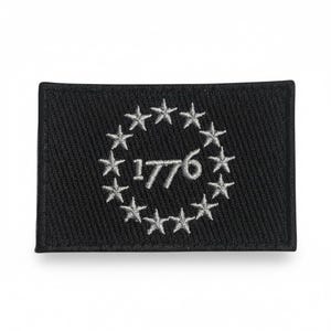 以下が含まれることがあります： 黒い長方形の刺繍ワッペン。中央に銀色の糸で「1776」という数字が刺繍されており、その周囲を13個の銀色の星が円形に囲んでいるデザインで、背景は無地の黒い布地で構成されています。