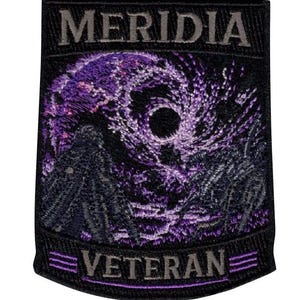 May include: A rectangular embroidered patch with a black background. The top features the text MERIDIA in grey. The center displays a swirling purple and black cosmic nebula design. The bottom features the text VETERAN in grey with purple lines.