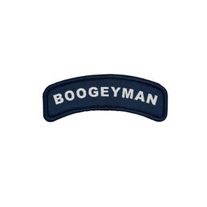 Può includere: Una toppa ricamata blu navy con la scritta "BOOGEYMAN" in bianco, arcuata nella parte superiore. La toppa ha una forma rettangolare curva ed è posta su uno sfondo bianco.