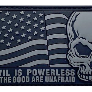 May include: A rectangular morale patch in shades of dark gray and light gray. The design features a stylized American flag, a skull on the right side, and the text "EVIL IS POWERLESS IF THE GOOD ARE UNAFRAID" embossed in capital letters at the bottom.