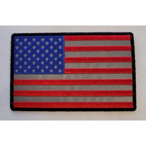 Puede incluir: Un parche rectangular bordado con el diseño de la bandera de los Estados Unidos. El cantón es azul con estrellas bordadas, mientras que el campo consta de franjas horizontales alternas de color rojo y gris. El parche tiene un borde negro.
