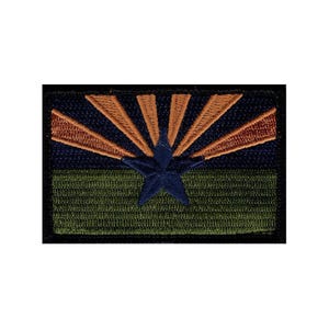 May include: A rectangular embroidered patch featuring a dark blue star centered over a horizontal split design. The top half displays radiating copper-colored sun rays against a dark blue background, while the bottom half is a solid olive green.