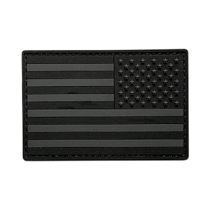 May include: A rectangular morale patch featuring a monochromatic black and dark grey American flag design. The patch displays raised horizontal stripes and a field of stars in the upper left corner, all rendered in a consistent dark colour scheme.